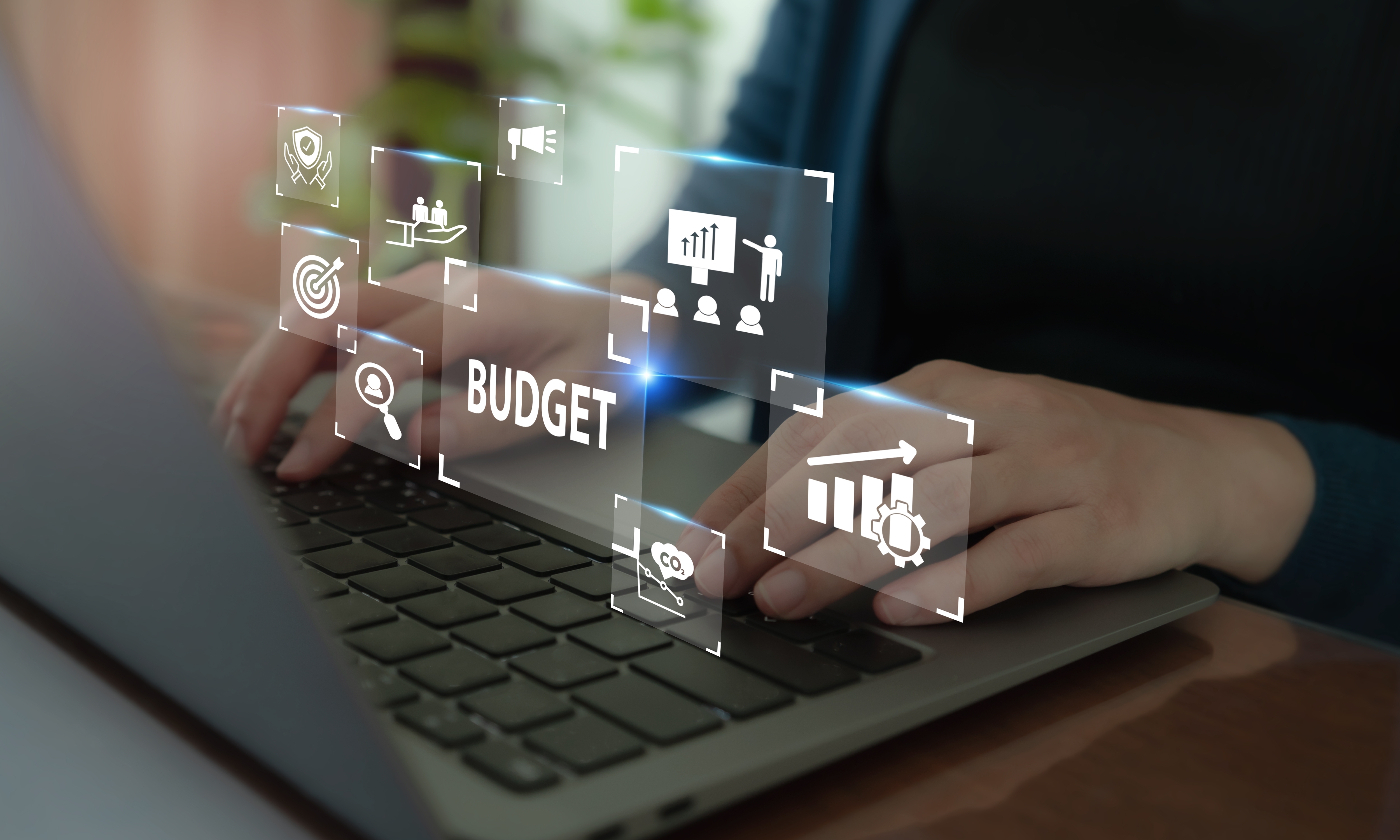 Hands typing on a laptop keyboard with digital overlays showing the word 'BUDGET' and icons representing business concepts like security, growth, optimization, and analysis.