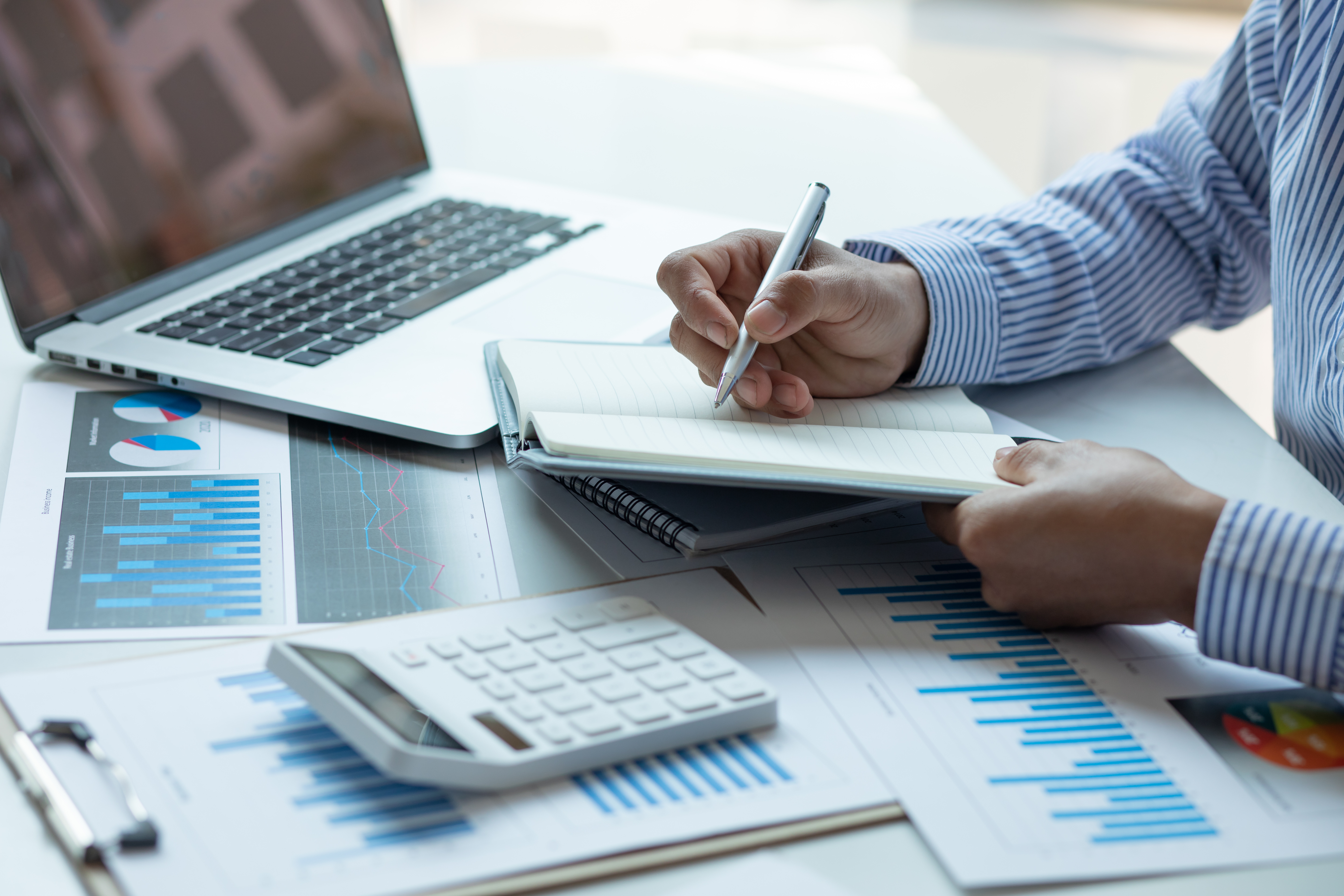 Close-up of a person writing in a notebook at a desk, surrounded by a laptop, a white calculator, and financial documents with charts and graphs, symbolizing accounting and financial analysis.