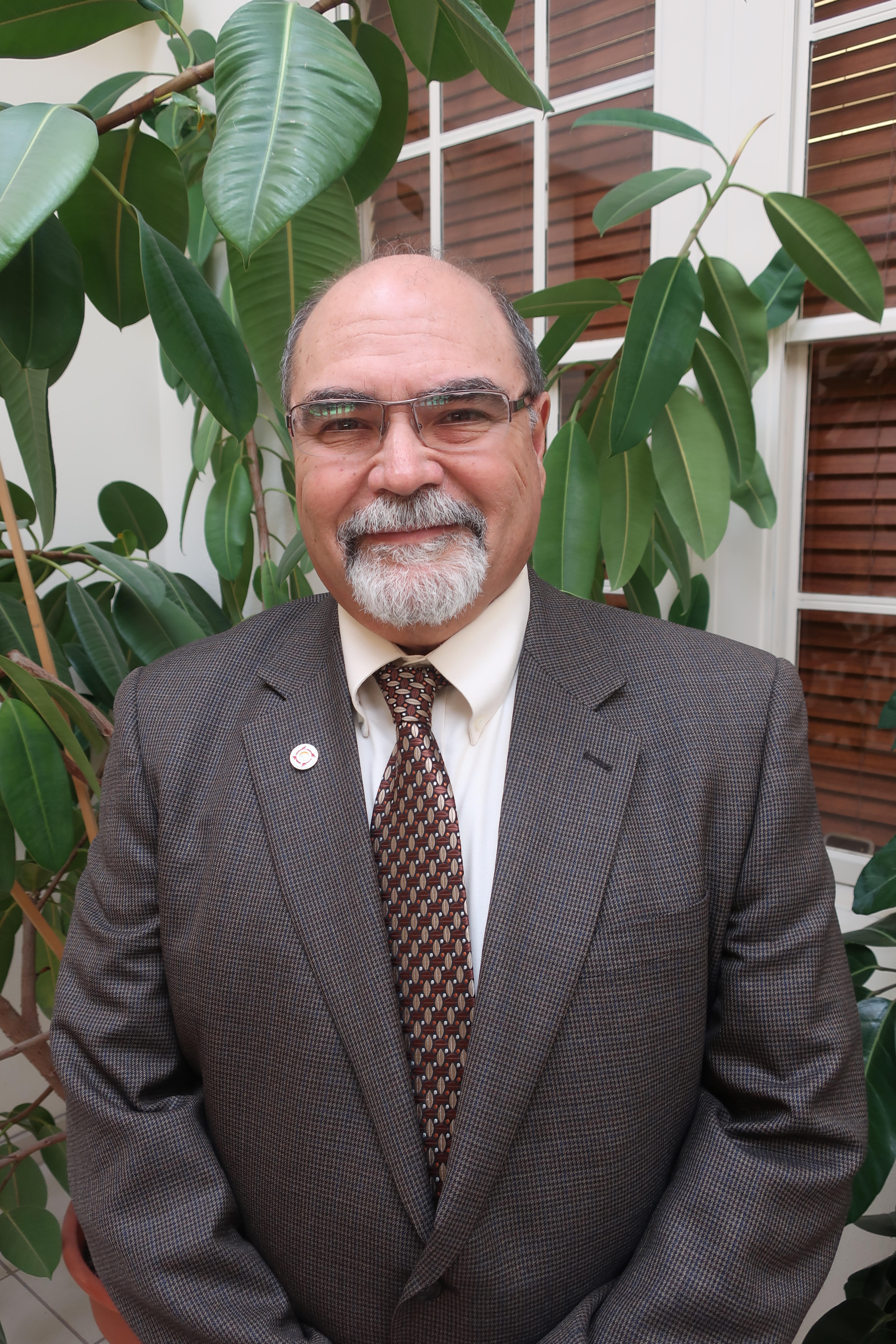 Mark Apodaca standing indoors in front of green plants and a window with wooden blinds, wearing a brown checked blazer, a white shirt, and a patterned tie. He is smiling confidently, and a small lapel pin is visible on his blazer.